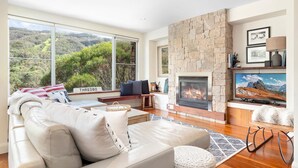 A modern living room with a stone fireplace, large windows overlooking a forested area, a comfortable sofa, and a flat-screen TV.