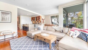 A spacious living room with a large sofa, a wooden coffee table, and a patterned rug. There is a kitchen area with wooden cabinets and a dining table visible in the background.