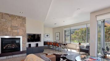 A spacious living area with a stone fireplace, a flat-screen TV, a dining area with chairs, and large windows offering a view of the outdoors.