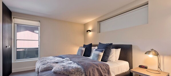 Un dormitorio con una cama, una mesita de noche con lámpara, una ventana y una luz empotrada en la pared.