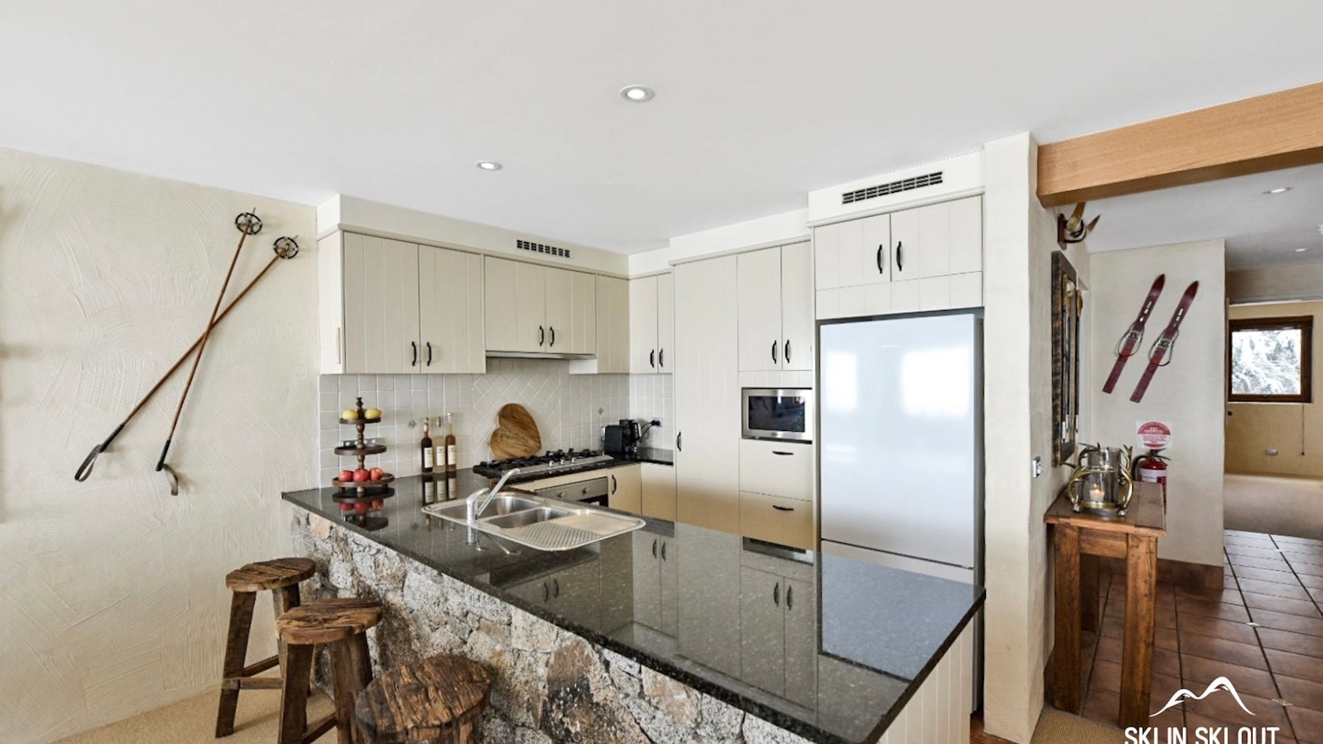 A modern kitchen with white cabinets, a marble island, and a built-in oven. There are wooden stools and a ski rack visible.