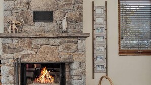 Une cheminée en pierre avec un feu de bois, un panier et une fenêtre avec des stores.