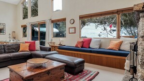 Un salon avec un canapé en cuir marron, une table basse en bois et un canapé bleu agrémenté de coussins. De grandes fenêtres sont ornées de rideaux, une horloge est accrochée au mur et une cheminée en pierre est présente.