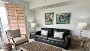 Una sala de estar con un sofá de cuero negro, dos mesitas de noche con lámparas, una mesa de centro de madera, un sillón beige y un ventanal con contraventanas de madera.
