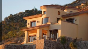A multi-story house with a stone facade, balconies, and a mountainous backdrop. - Cabos Montecristo Villa w/ unobstructed breathtaking ocean view (Cabo San Lucas)