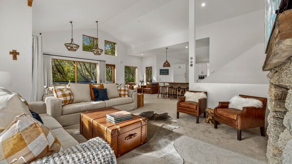 A spacious living room with a large sofa, leather armchairs, and a coffee table. There are large windows with a view of trees, and a kitchen area is visible in the background. - Ski In Ski Out #01 (Ski-In-Ski-Out)