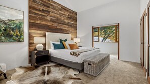A bedroom with a wooden wall, a large window, a bed with multiple pillows, a nightstand, and a fur rug. - Ski In Ski Out #01 (Ski-In-Ski-Out)