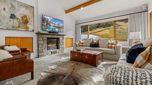 A living room with a stone fireplace, a leather sofa, a large window with a view of a landscape, and a television mounted on the wall. - Ski In Ski Out #01 (Ski-In-Ski-Out)