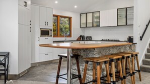 A modern kitchen with white cabinetry, a wooden island with bar stools, a built-in oven, and a window with a view of greenery. - Ski In Ski Out #01 (Ski-In-Ski-Out)