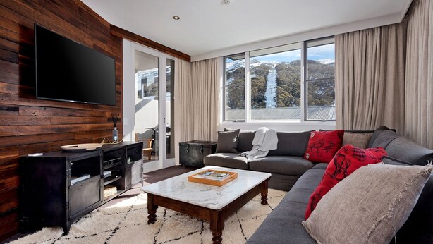 A modern living room with a flat-screen TV mounted on a wooden wall, a grey sofa with red and black cushions, a wooden coffee table, and a large window offering a view of a snowy landscape. - KoKo Nanook - Squatters 15 (Central Village)