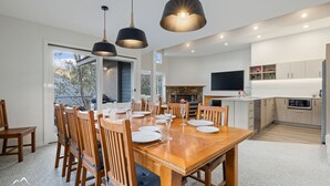 A modern dining area with a wooden table set for six, a stone fireplace, and a kitchen with white cabinets and a flat-screen TV.