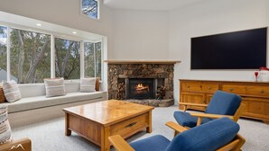 A living room with a stone fireplace, a large window with a view of trees, a wooden coffee table, and blue armchairs.