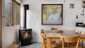 Een gezellige kamer met een houtkachel, een eettafel met stoelen en een raam met uitzicht op een huis.