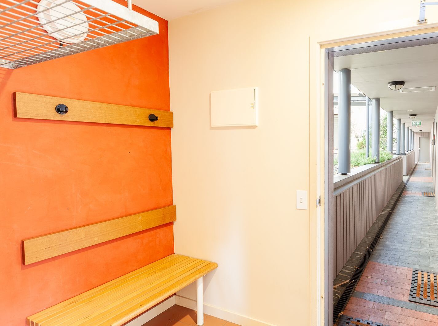 A hallway with an orange wall, a wooden bench, and a white door leading to a covered area with columns and a radiator.