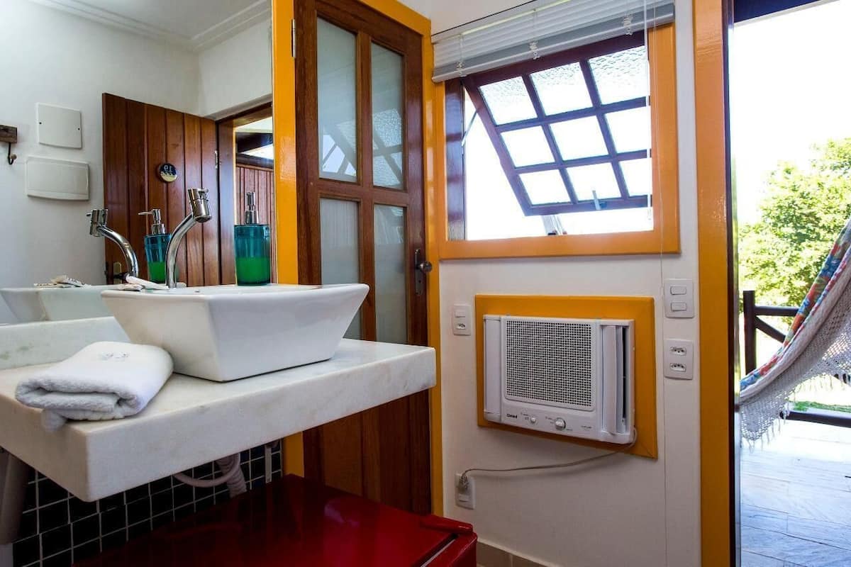 Ein kleines Badezimmer mit einem weißen Waschbecken, zwei Wasserhähnen und einem grünen Seifenspender. Ein Fenster mit Holzrahmen und eine weiße Klimaanlage, die an der Wand befestigt ist.