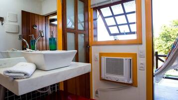 Een compacte badkamer met een witte wastafel, twee kraanwerk en een groene zeepdispenser. Er is een raam met houten kader en een witte airconditioningunit aan de muur bevestigd.