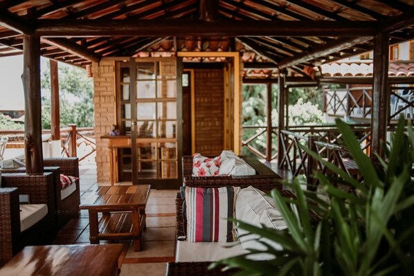 A covered outdoor seating area with wicker furniture, a wooden table, and a view of the garden. - Pousada Janellas do Mar (Búzios)