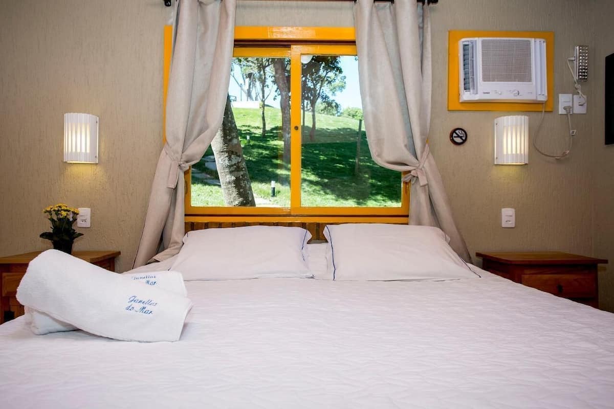 Ein Hotelzimmer mit einem Bett, zwei Nachttischen, einem Fenster mit Vorhängen, einer Wand-Klimaanlage und einer Wanduhr.