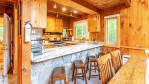 Ein Holzblockhaus-Interieur mit einer Steintheke, hölzernen Barhocker, einer Küche mit modernen Geräten und großen Fenstern.