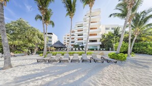 A beachside hotel with sun loungers, palm trees, and a modern building with balconies. - 3BR Oceanfront Condo in Sac Bajo (Isla Mujeres)