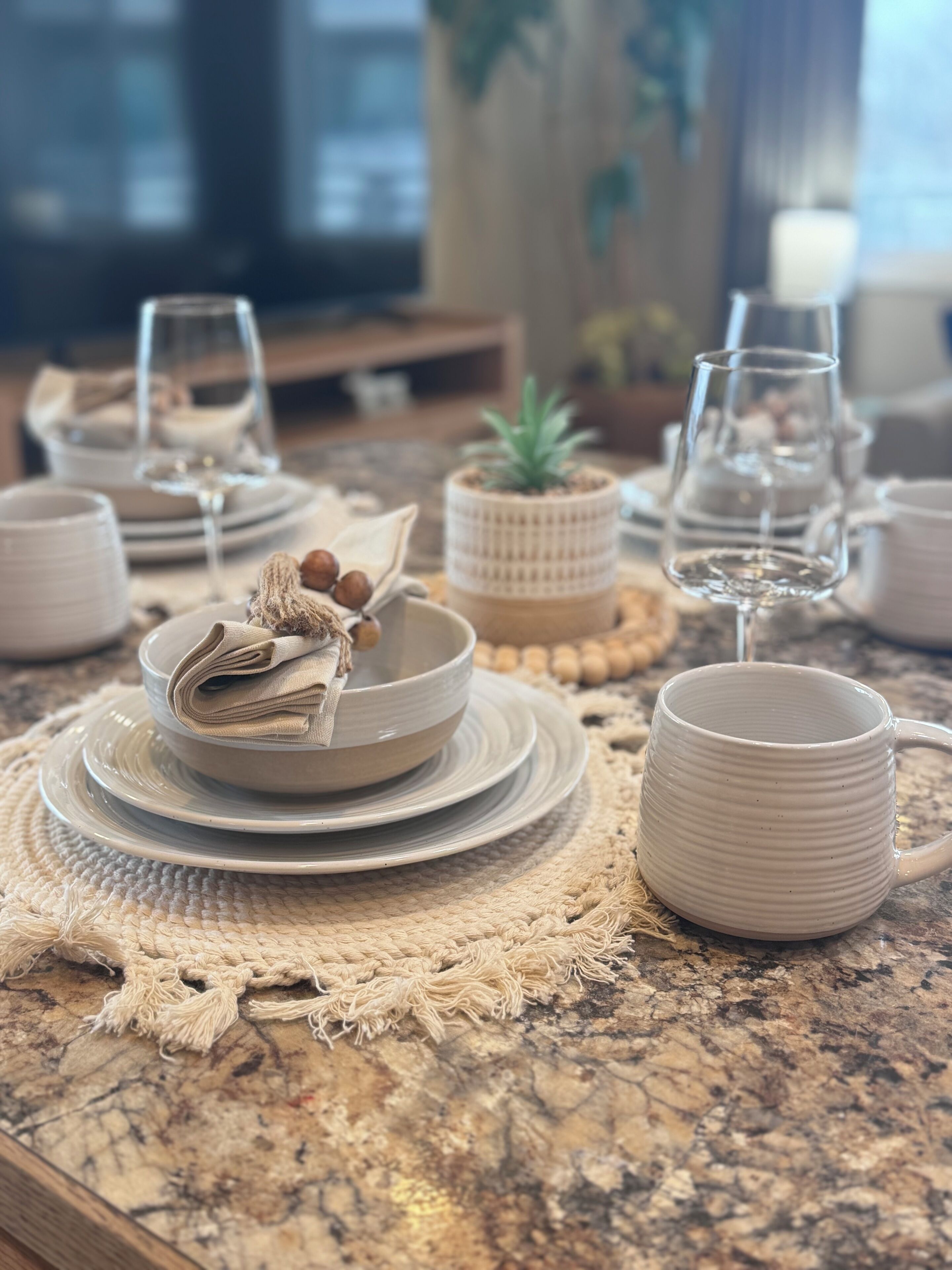 Una mesa de comedor dispuesta con platos, tazones y una taza, con un mantel tejido y una pequeña planta en maceta.