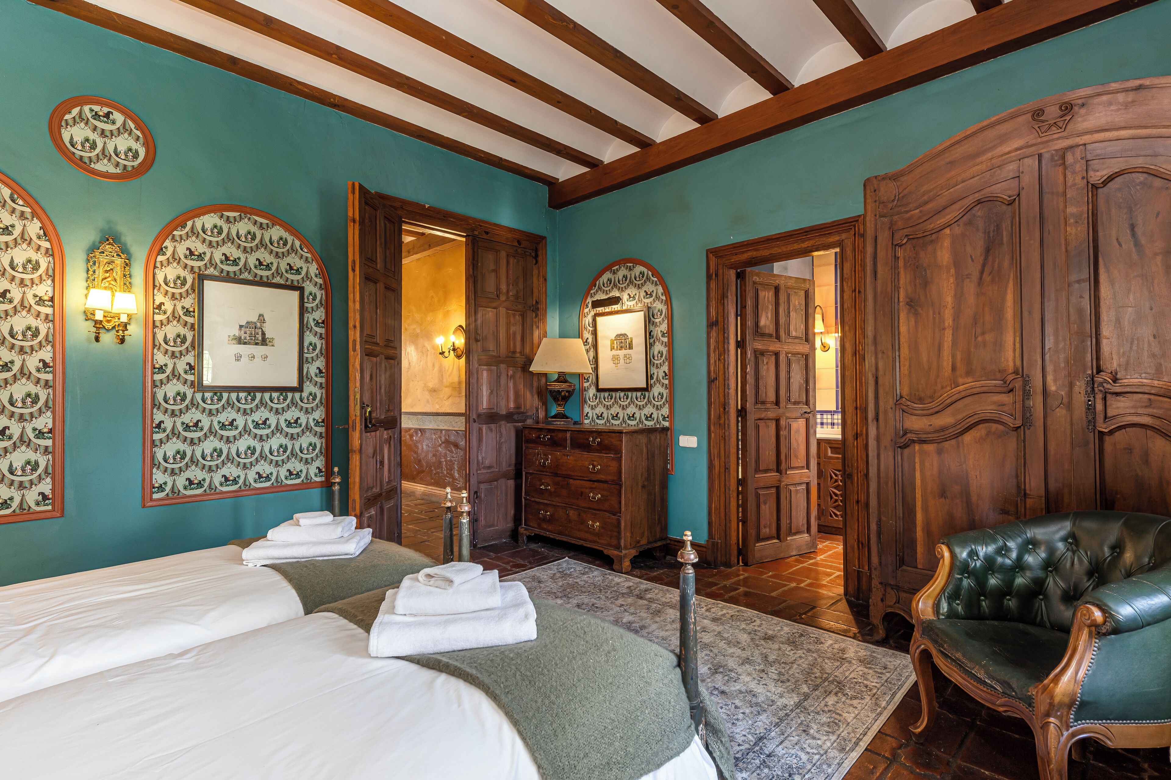 A hotel room with a large bed, a green armchair, wooden wardrobe, and a mirror with a framed picture.