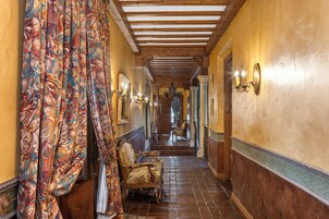 A hallway with patterned curtains, wooden chairs, and wall sconces.