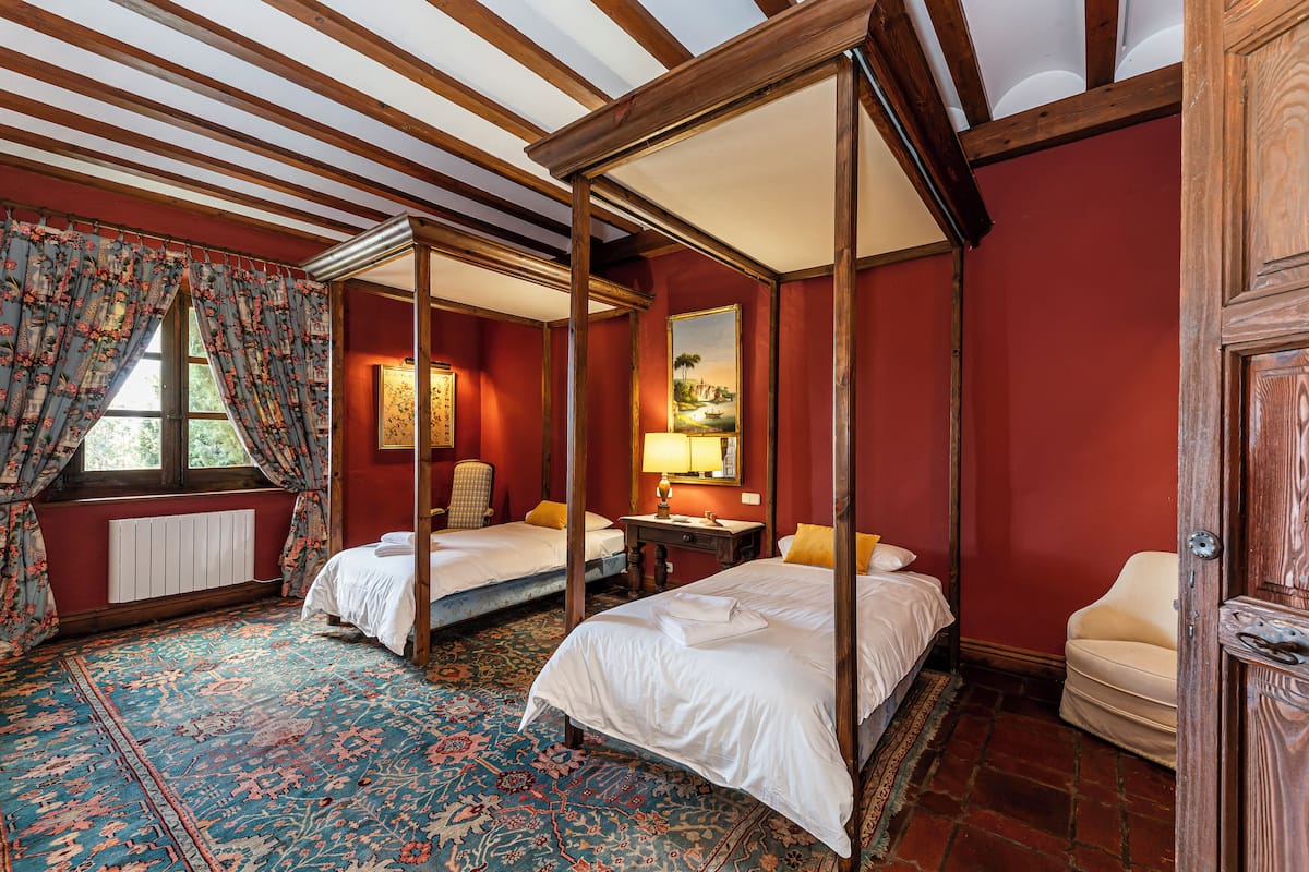 A four-poster bed, two single beds, a chair, a table, and a radiator in a room with red walls and a patterned carpet.