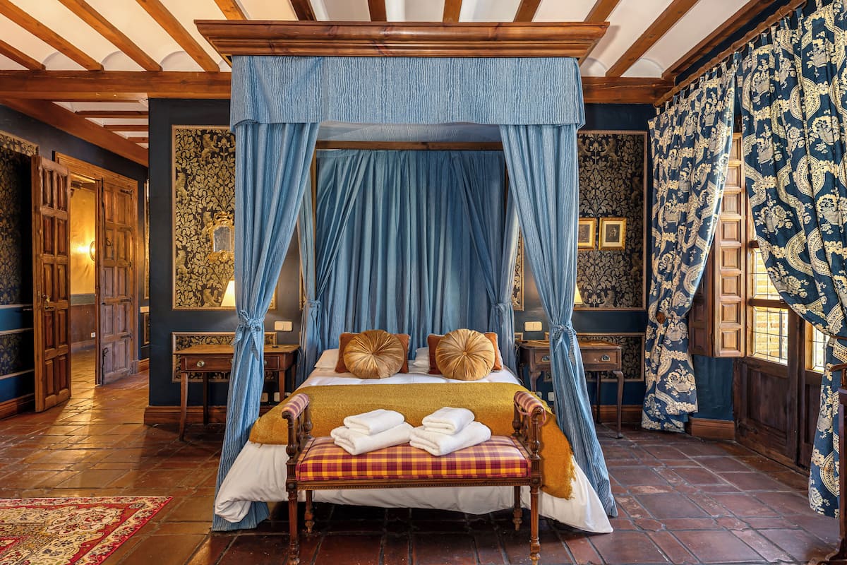 A traditional bedroom with a four-poster bed, a bench with towels, and ornate curtains.