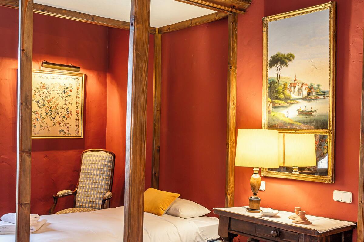 A room with a four-poster bed, a single bed, a chair, a bedside table with a lamp, and a painting on the wall.