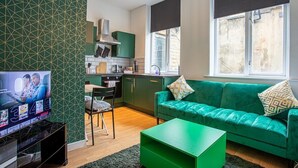 A modern living room with a green sofa, a flat-screen TV, and a kitchen in the background. - Blissfully Peaceful 1 Bed Flat In Crown Street (West Yorkshire)