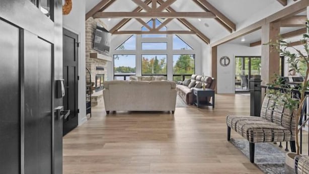 A spacious living area with a high ceiling and exposed beams, a central seating area, and large windows offering a view of the outdoors.