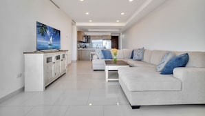 A modern living room with a sofa, coffee table, and TV stand. There is a kitchen in the background.
