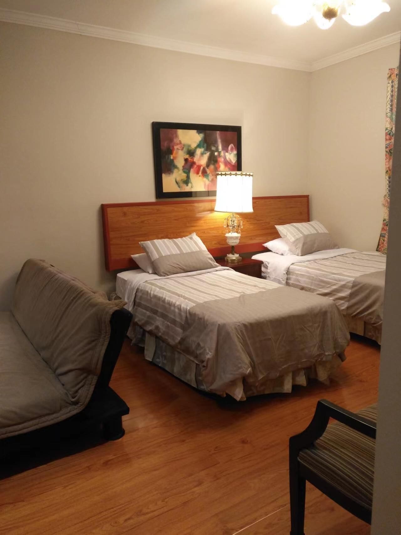 A hotel room with two beds, a wooden headboard, a nightstand with a lamp, and a colorful abstract painting on the wall.