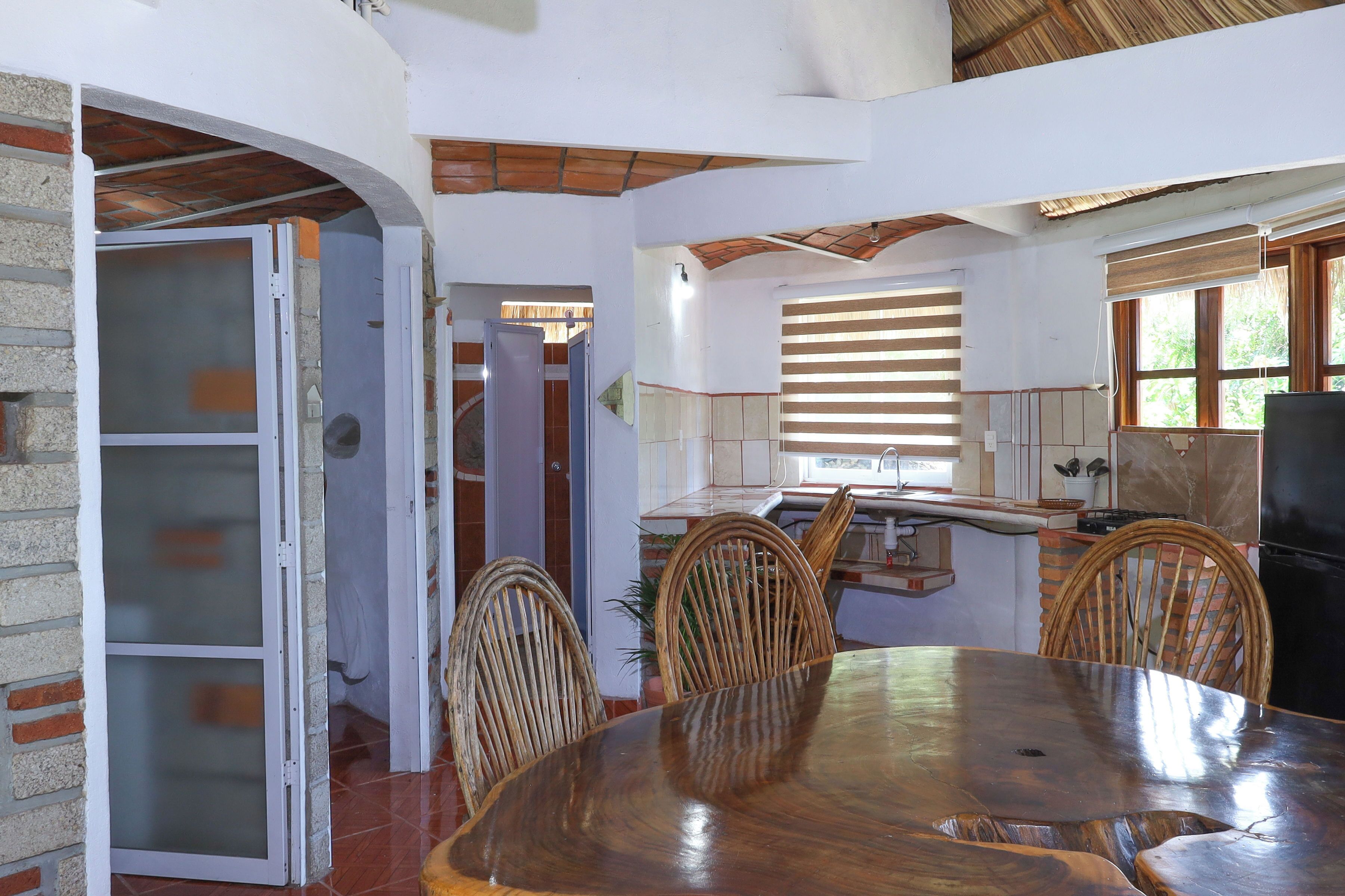 A dining area with a wooden table and chairs, a kitchen in the background, and a view of the outdoors through a window.