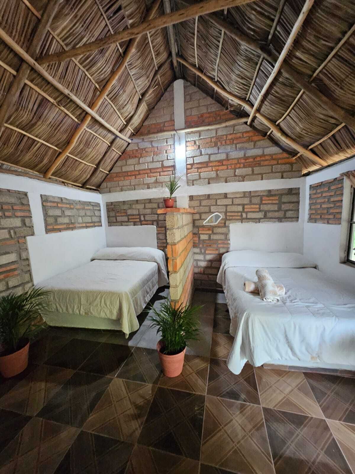 A room with two beds, a brick wall, a thatched roof, and potted plants.