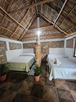 A room with two beds, a brick wall, a thatched roof, and potted plants.