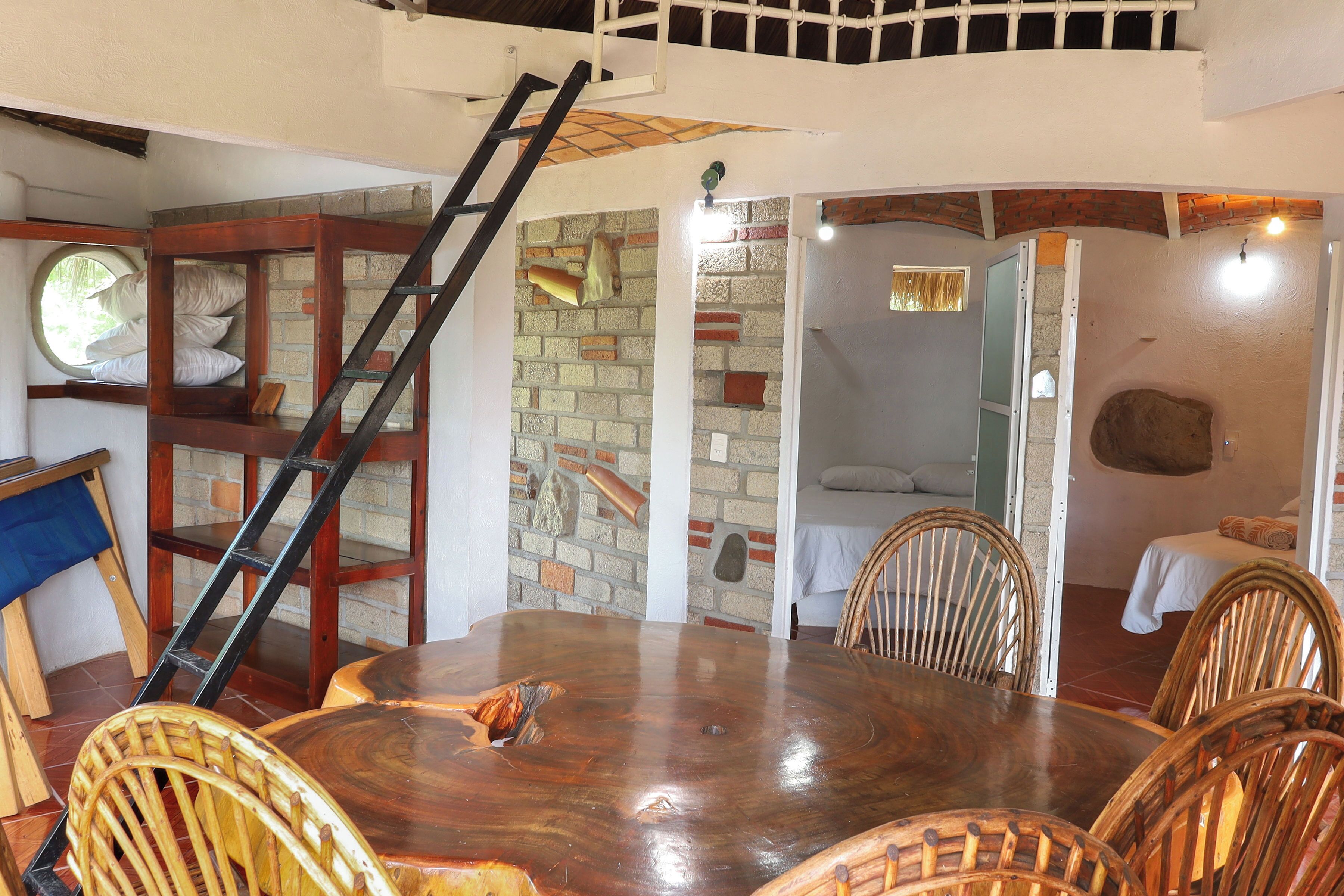 A dining area with a wooden table and chairs, a brick wall, and a loft bedroom with a bed and window.