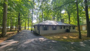 Une petite cabane d’un seul étage, avec un toit en métal, entourée de grands arbres et bordée d’une allée de gravier.