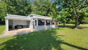 A covered patio with a grill and chairs, a small deck with American flag decorations, and a grassy yard with trees. - 20% Off Lakefront Tiny Bird Dock, Hot Tub, Firepit, Boat House, ParkLike Setting (Dandridge)