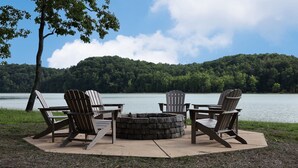 A fire pit with four Adirondack chairs around it, set on a concrete platform by a lake with a forested hill in the background. - 20% Off Lakefront Tiny Bird Dock, Hot Tub, Firepit, Boat House, ParkLike Setting (Dandridge)