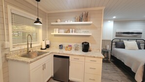 A compact kitchen with a sink, dishwasher, and a coffee maker. There is a bed with a view of a landscape painting. - 20% Off Lakefront Tiny Bird Dock, Hot Tub, Firepit, Boat House, ParkLike Setting (Dandridge)