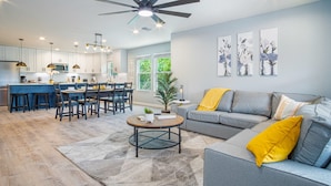 A modern living room with a grey sofa, yellow cushions, a coffee table, and a dining area with chairs and a kitchen in the background. - Comfortable 4BR 2 5BA Family Home Near Riverwalk (San Antonio)
