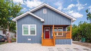 A small, single-story building with a red door, a wooden front porch, and a gravel driveway. - Comfortable 4BR 2 5BA Family Home Near Riverwalk (San Antonio)