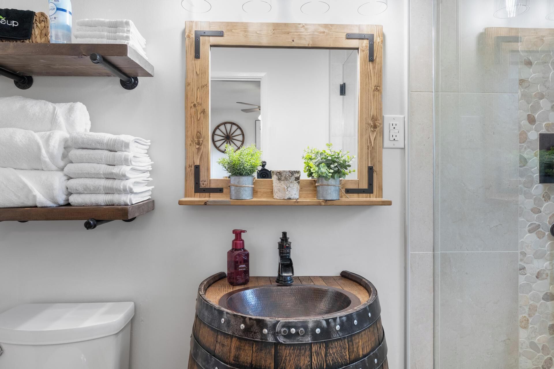 A bathroom with a wooden barrel sink, a wooden-framed mirror, and a shelf with towels and potted plants.