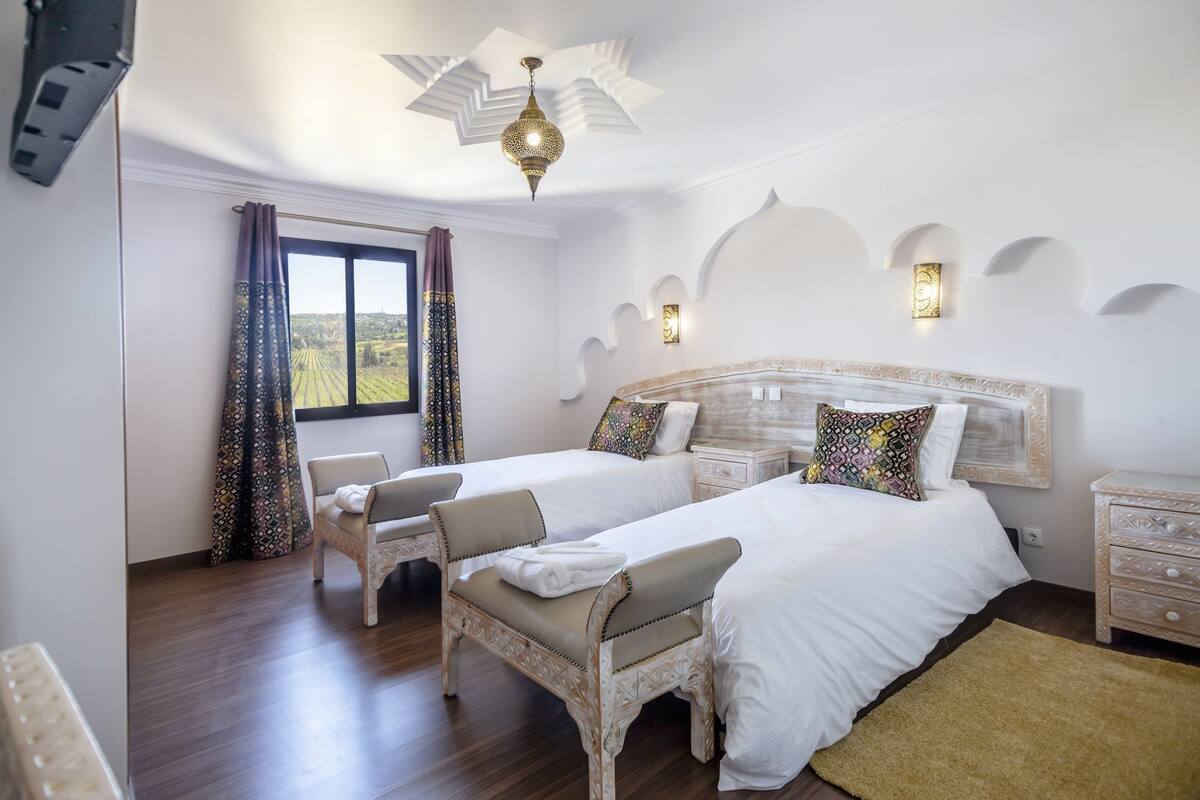 A bedroom with two beds, a window with curtains, a chandelier, and a nightstand.