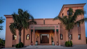 A building with a traditional architectural style, featuring a central entrance with a decorative archway and a large wooden door. There are palm trees and potted plants around the entrance, and the building is surrounded by a paved area. - Al Karob Yoga Boutique Hotel (Albufeira)