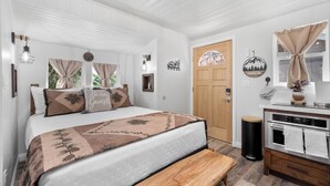 A bedroom with a bed, a bench, a wooden door, a kitchen area, and a window with curtains. - Julian Cafe Couples Cottage - Breakfast Included at The Julian Cafe & Bakery! (Julian)