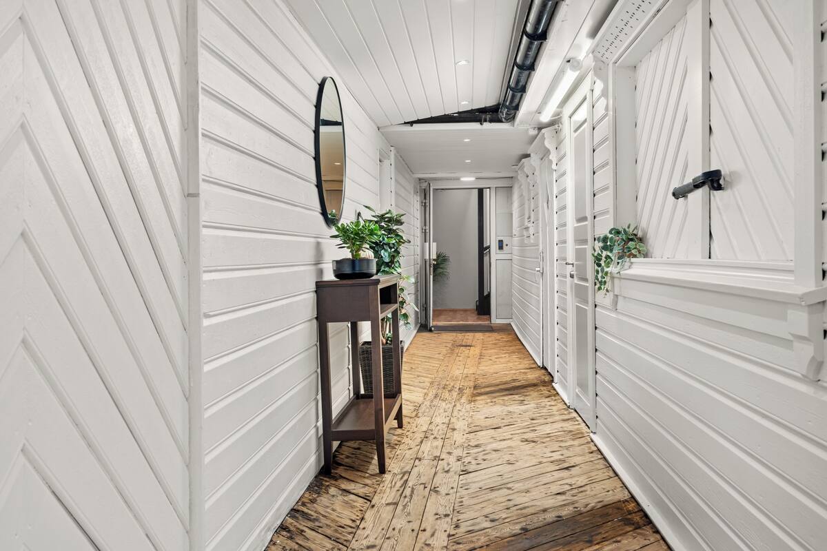 Un couloir aux murs recouverts de lambris blancs, un plancher en bois, un miroir, une petite table avec une plante et une porte dotée d’une poignée.
