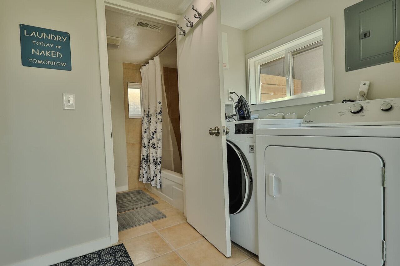 A laundry room with a sign reading "Laundry today or naked tomorrow," a washing machine, a dryer, a window, and a shower area with a curtain.
